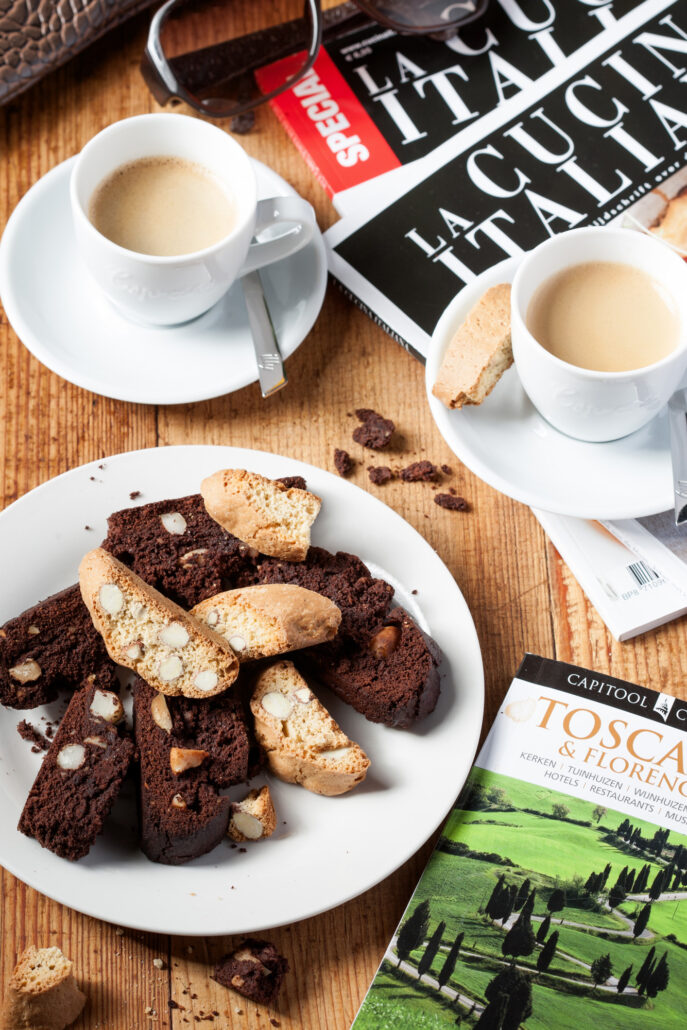 Chocolade biscotti en amandel cantuccini op wit bord met espresso en Italiaanse tijdschriften op tafel.
