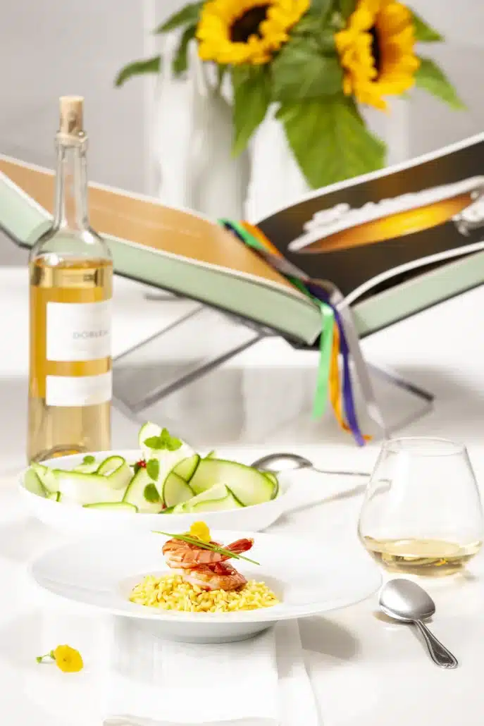 Risotto met saffraan en garnalen geserveerd op een wit bord met een glas witte wijn en een frisse courgettesalade op de achtergrond.