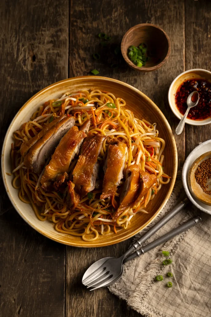 Chow Mein noedels met knapperige groenten en geroosterde eend, geserveerd in een schaal met chili-olie en sesamzaad op houten tafel.